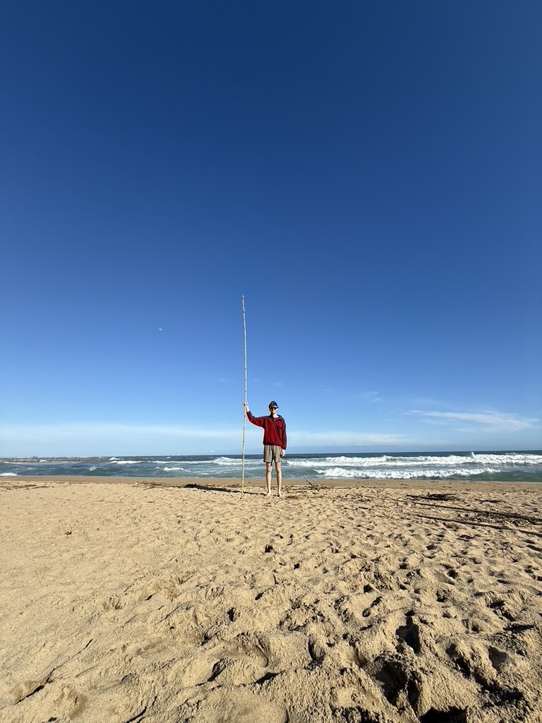 Gunner standing on a beach holding a tall pole beside the ocean.